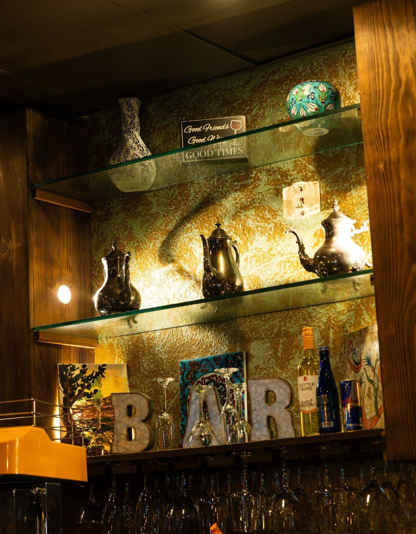 The back bar at KZ Lounge — silver teapots and painted ceramics on lit glass shelves above marquee BAR letters and rows of hanging glasses