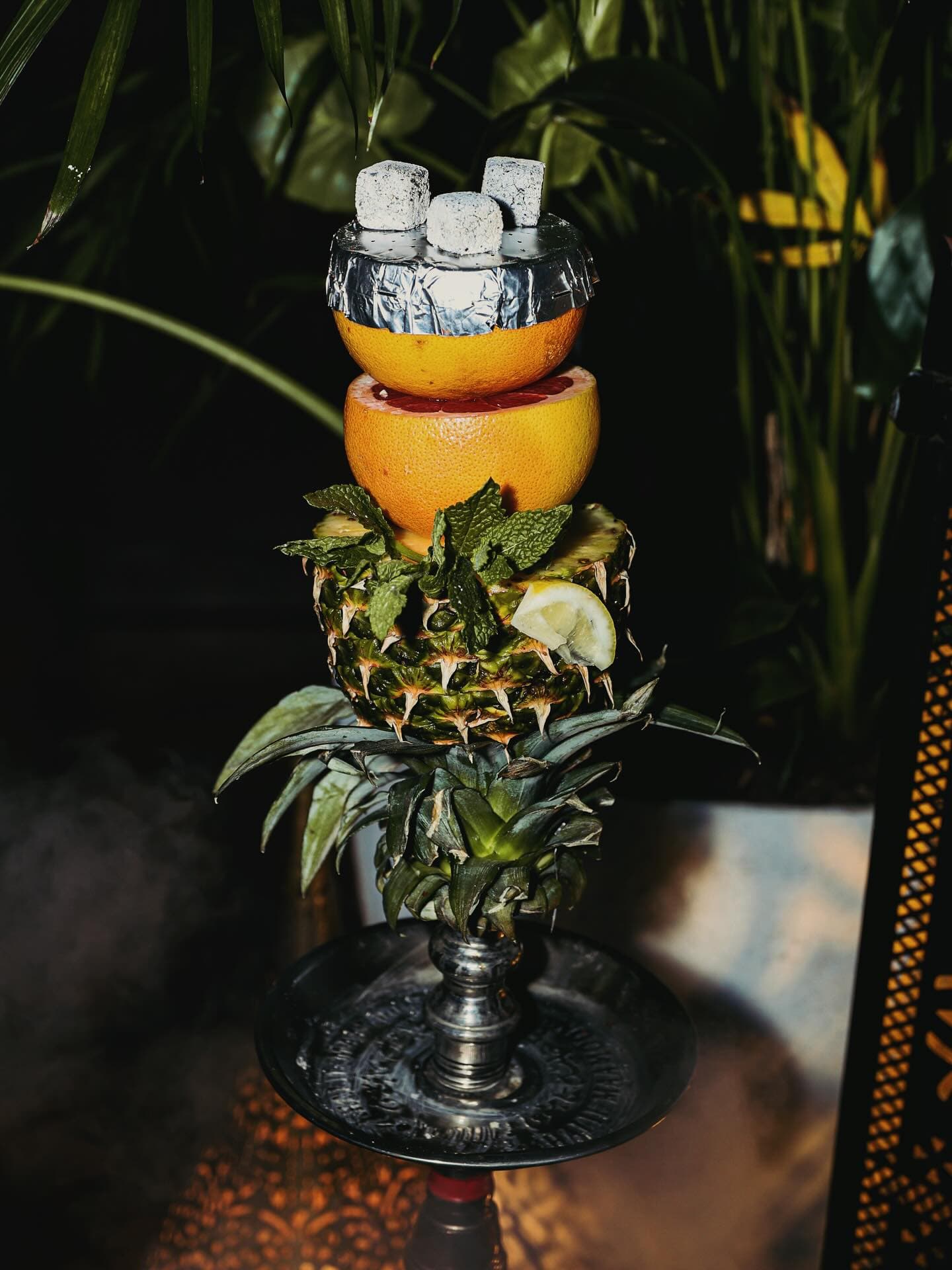 A hookah head built from a whole pineapple and grapefruit, coals lit, mint tucked between the fruit